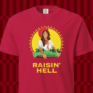 May include: A red short-sleeved t-shirt featuring a graphic of a person in a red bonnet and white top holding a basket of green grapes. Behind the person is a yellow sunburst design. The text RAISIN' HELL is printed in yellow below the graphic.