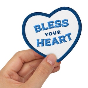 Może przedstawiać: Naszywka w kształcie serca z białym środkiem i niebieską obwódką. Słowa "BLESS YOUR HEART" są wyhaftowane na niebiesko pośrodku. Naszywka jest trzymana w dłoni osoby na białym tle.