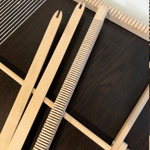 May include: A wooden weaving loom with white threads and several wooden tools. The loom is rectangular with a series of notches along the top and bottom edges. Three wooden tools are visible, including a ridged beater and two shuttles.