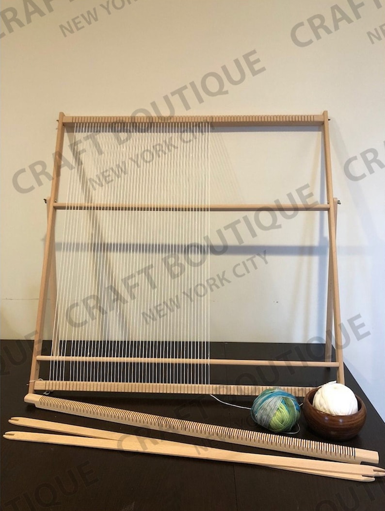 May include: A wooden weaving loom with white warp threads, along with weaving tools and yarn. The loom is rectangular, with a frame and horizontal bars. Two balls of yarn, one white and one multicolored, are also present. The text "CRAFT BOUTIQUE NEW YORK CITY" is visible.