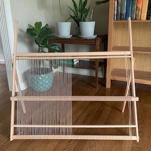 May include: A wooden weaving loom with vertical threads stretched across its frame. The loom is light-colored, with a simple design. The threads are a light beige color. The loom is set up in a room with plants and a bookshelf.