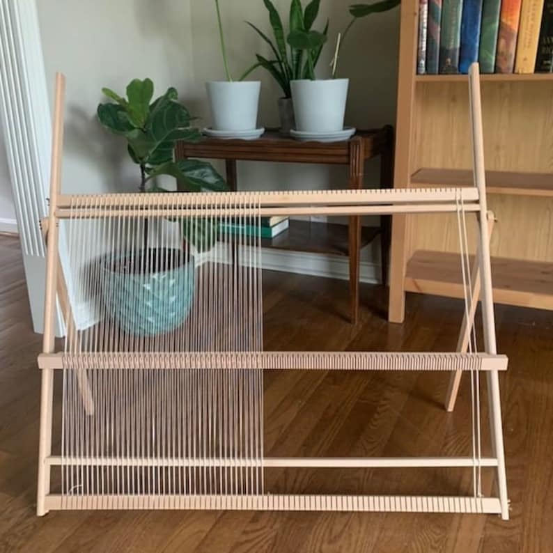 May include: A wooden weaving loom with vertical threads stretched across its frame. The loom is light-colored, with a simple design. The threads are a light beige color. The loom is set up in a room with plants and a bookshelf.