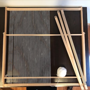 May include: A wooden weaving loom with white yarn strung across the frame. Two wooden weaving tools and a ball of white yarn are next to the loom. The loom is on a dark brown surface.
