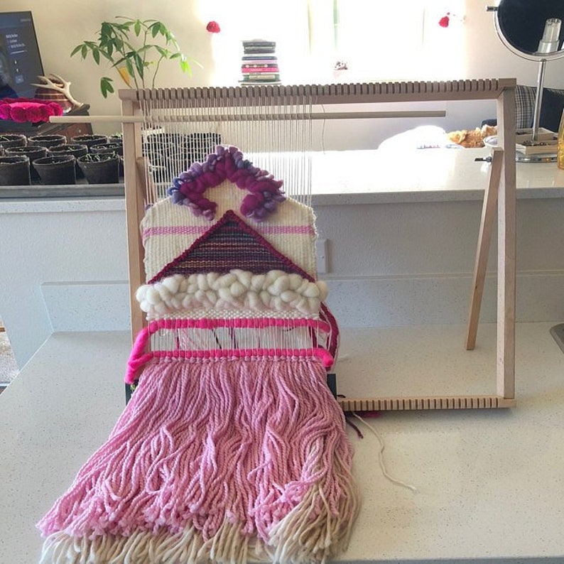 May include: A pink and white woven wall hanging with a geometric design, hanging on a wooden loom.