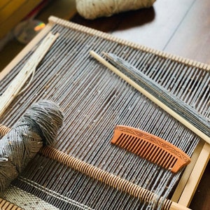 May include: A wooden loom with gray yarn wound around it. A wooden comb and a wooden dowel are also on the loom.