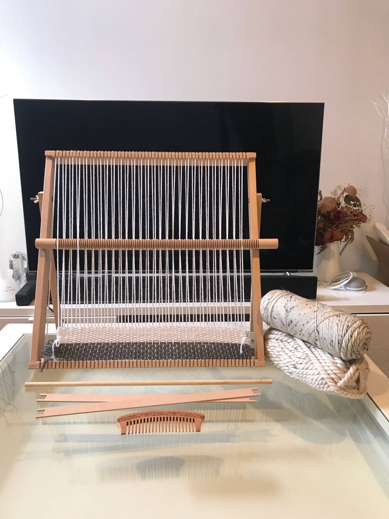 May include: A wooden loom with white yarn strung across it, a wooden comb, two wooden weaving needles, and two balls of white yarn on a glass table.