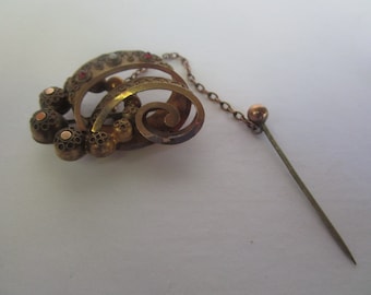 May include: A gold-toned brooch with a decorative design featuring a swirl and multiple round accents. The brooch has a pin and chain attached.