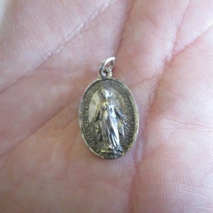 May include: Silver oval pendant featuring a depiction of the Virgin Mary with a crown and a blue robe. The pendant has a detailed design and a smooth finish.