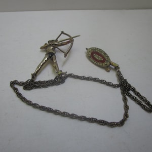 May include: A gold chain necklace with two pendants. One pendant is a figure of an archer with a bow and arrow. The other pendant is a round, red and white enamel design.