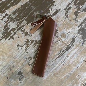 May include: A brown leather pen sleeve featuring white stitching along the edges, a metal lobster clasp at the top, and a small leather tag with the word JUNG printed in white. The sleeve is designed to hold a single writing instrument securely.