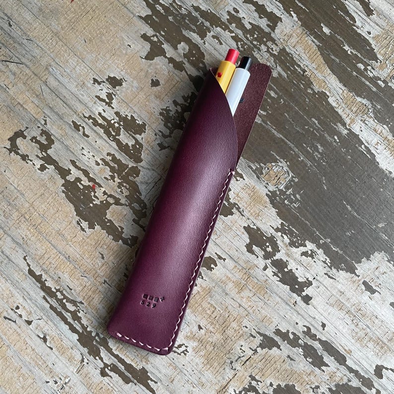 May include: A deep burgundy leather pen sleeve featuring white contrast stitching along the edges and a small embossed logo near the bottom. Two pens, one with a yellow barrel and one with a white barrel, are inserted into the top opening.