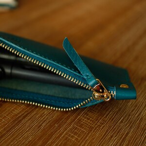 May include: Teal leather pen case with a gold zip. The zip is partially open, revealing the interior. The case has a small leather pull tab. The case is on a wooden surface.
