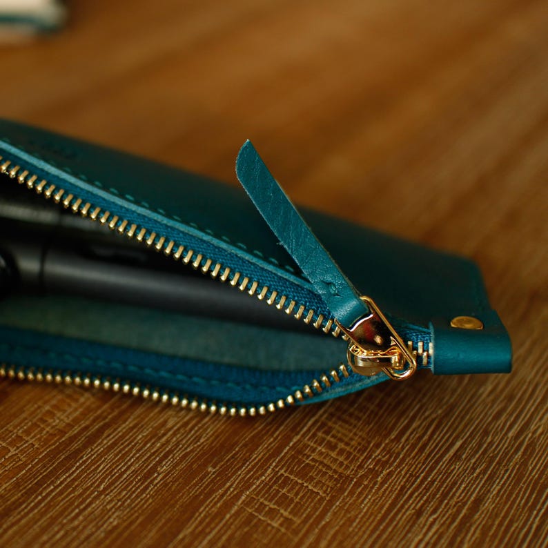May include: Teal leather pen case with a gold zip. The zip is partially open, revealing the interior. The case has a small leather pull tab. The case is on a wooden surface.