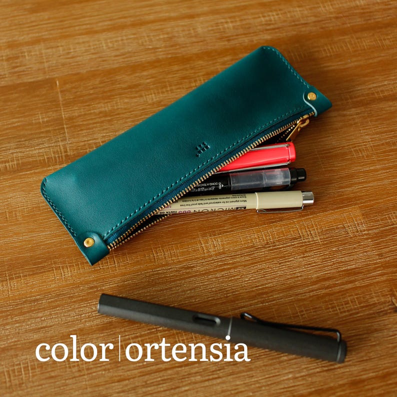 May include: Teal leather pencil case with a gold zip, containing pens and markers. The case has a small embossed logo and is partially open. A black pen rests on the wooden surface. The text "color ortensia" is at the bottom.