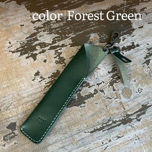 May include: A forest green leather pen sleeve featuring white contrast stitching, a metal swivel clasp, and a small leather tag with the text JUNG. The sleeve has a diagonal opening at the top and a small embossed logo near the bottom edge.