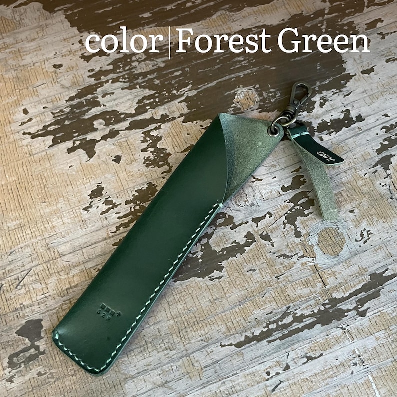 May include: A forest green leather pen sleeve featuring white contrast stitching, a metal swivel clasp, and a small leather tag with the text JUNG. The sleeve has a diagonal opening at the top and a small embossed logo near the bottom edge.