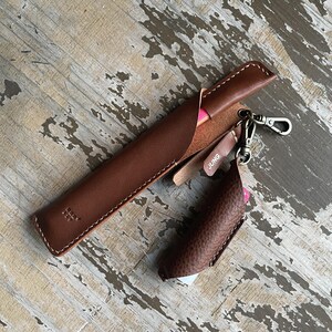 May include: A smooth brown leather pen case with white stitching and a small embossed logo, paired with a textured brown leather lighter sleeve attached by a metal swivel clasp. A small leather tag with the word JUNG is attached to the clasp.