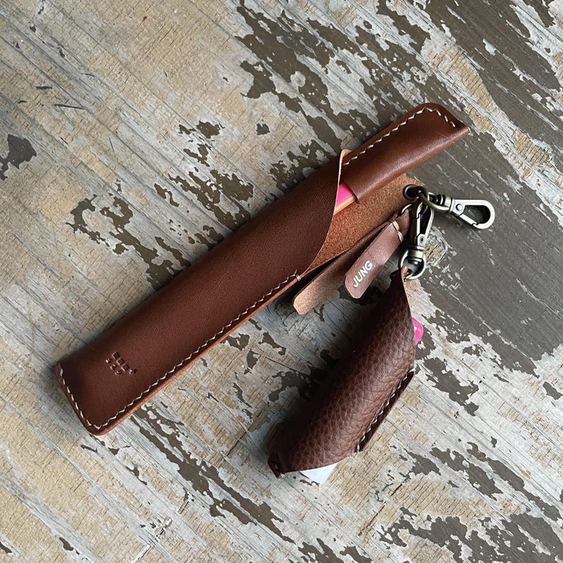 May include: A smooth brown leather pen case with white stitching and a small embossed logo, paired with a textured brown leather lighter sleeve attached by a metal swivel clasp. A small leather tag with the word JUNG is attached to the clasp.