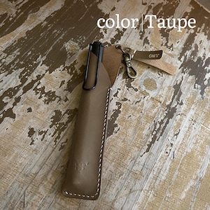 May include: A taupe leather pen sleeve containing a black pen with a clip. The sleeve features white stitching, a metal lobster clasp, and a small leather tag with the text JUNG. The image includes the text color | Taupe at the top.