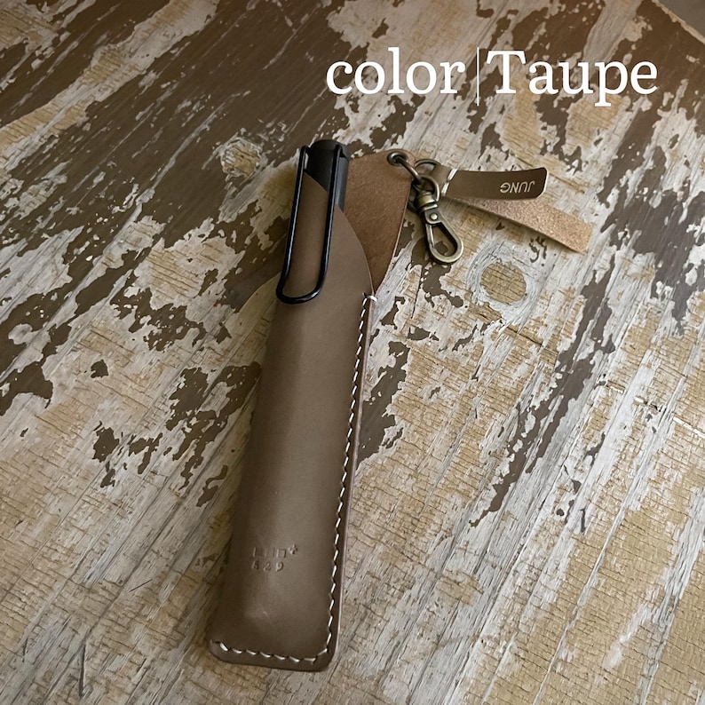 May include: A taupe leather pen sleeve containing a black pen with a clip. The sleeve features white stitching, a metal lobster clasp, and a small leather tag with the text JUNG. The image includes the text color | Taupe at the top.