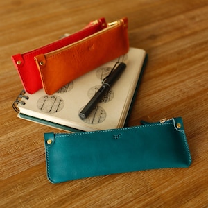 May include: Three leather pencil cases in red, orange, and teal, each with a gold zip and a small gold rivet. A black pen and a notebook with a spiral binding are also visible on a wooden surface.
