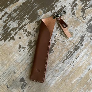 May include: A brown leather pen sleeve with a curved top opening, white contrast stitching, and a small metal lobster clasp attached to a leather tab. The tab features the text JUNG embossed in white. A small square logo is embossed on the front.