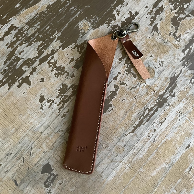 May include: A brown leather pen sleeve with a curved top opening, white contrast stitching, and a small metal lobster clasp attached to a leather tab. The tab features the text JUNG embossed in white. A small square logo is embossed on the front.