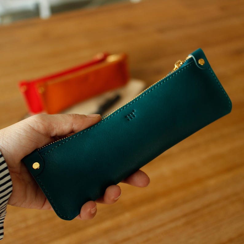 May include: Teal leather pencil case with a gold zip and gold rivets. The rectangular case has a small embossed logo and is held in a hand. A red and orange pencil case are in the background.