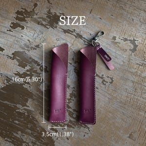 May include: Two purple leather pen sleeves with white stitching. One sleeve features a metal lobster clasp and a small leather tag with the text JUNG. The sleeves measure 6.30 inches in height and 1.38 inches in width as indicated by the text.