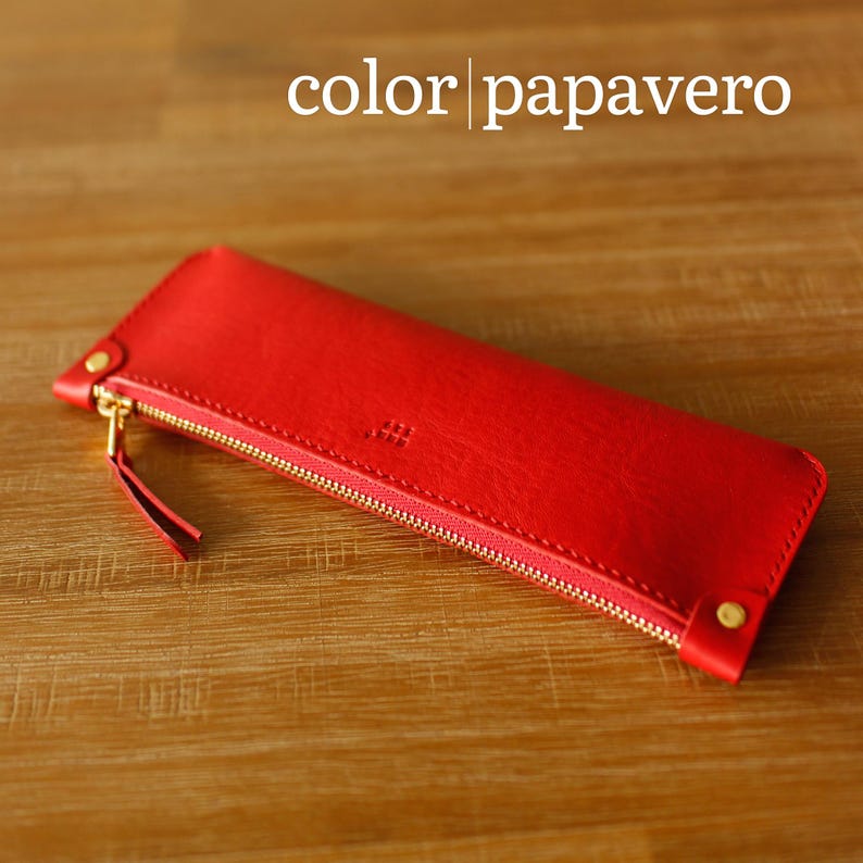 May include: A bright red leather pencil case with a gold zip and a small red pull tab. The rectangular case is closed with a gold zip. The text "color | papavero" is displayed at the top. The case is made of leather.