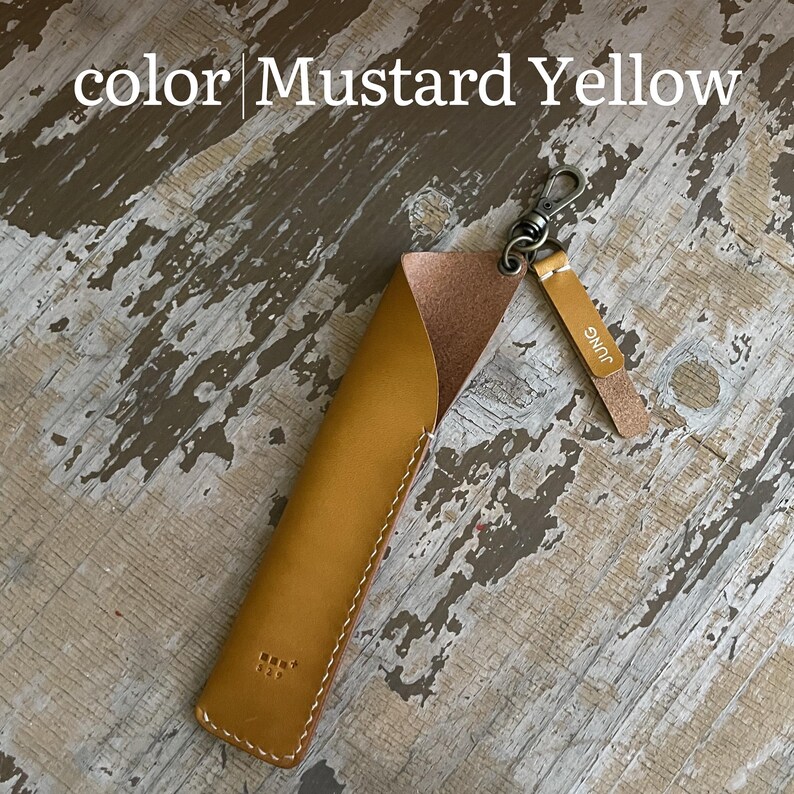May include: A mustard yellow leather pen sleeve with a matching leather tag featuring the embossed text JUNG. The sleeve includes a bronze-toned metal lobster clasp attached to a ring, and a small embossed logo is visible on the lower front surface.