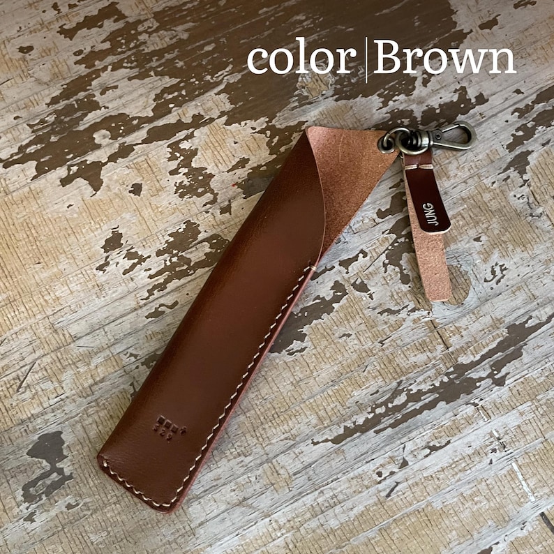 May include: A brown leather pen sleeve with white stitching, featuring a metal swivel clasp attached to a small leather tab embossed with the text JUNG. The sleeve has a diagonal opening and a small embossed logo near the bottom corner.