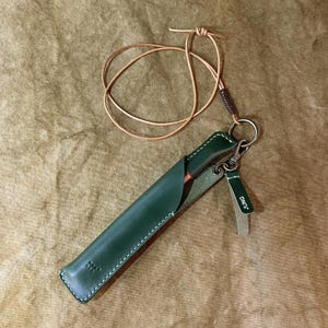 May include: A dark green leather pen sleeve with white stitching, featuring a long thin leather cord for wearing around the neck. A small matching green leather tag attached to a metal ring is embossed with the white text JUNG.