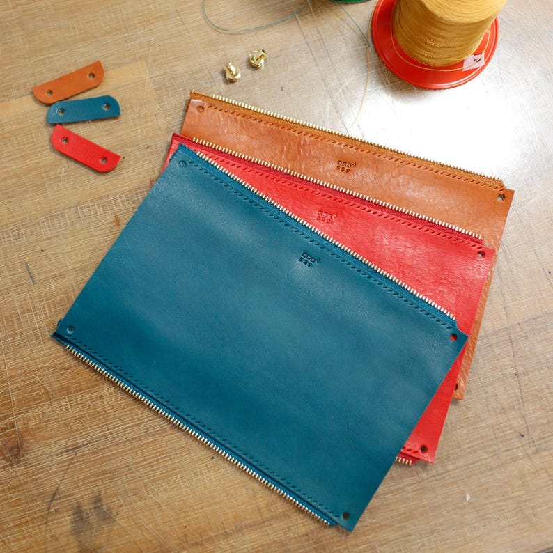 May include: Three leather zipper pouches in teal, red, and brown, stacked on a wooden surface. Each pouch has a gold zip and the brand name embossed on the front. Small leather tabs in orange, teal, and red are also visible.
