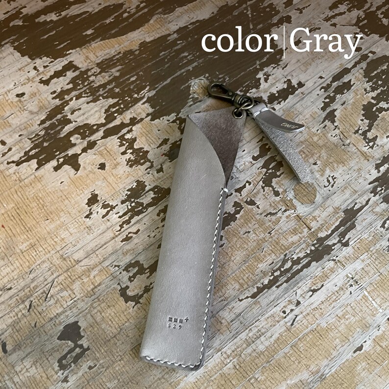 May include: A gray leather pen case featuring a diagonal opening, white contrast stitching, and a small metal lobster clasp attached to a leather pull tab. The text "color Gray" is visible at the top, and a small logo is embossed at the bottom.