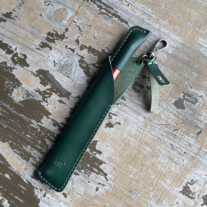 May include: A dark green leather pen sleeve with visible yellow stitching, containing a red pen. A small leather tag attached to a metal lobster clasp features the embossed text JUNG. The sleeve has a smooth finish and a pointed top edge.
