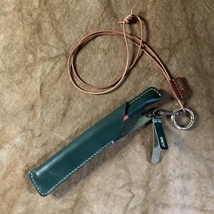 May include: A dark green leather pen sleeve with white stitching, featuring a long thin leather cord for carrying. A small dark green leather tag attached to a silver metal ring displays the word JUNG in white capital letters.