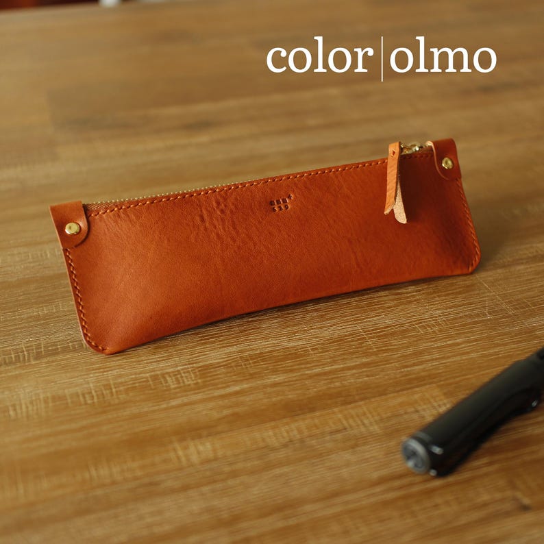 May include: An orange-brown leather pencil case with a gold zip and leather pull tab. The text "color olmo" is at the top. The pencil case is on a wooden surface.