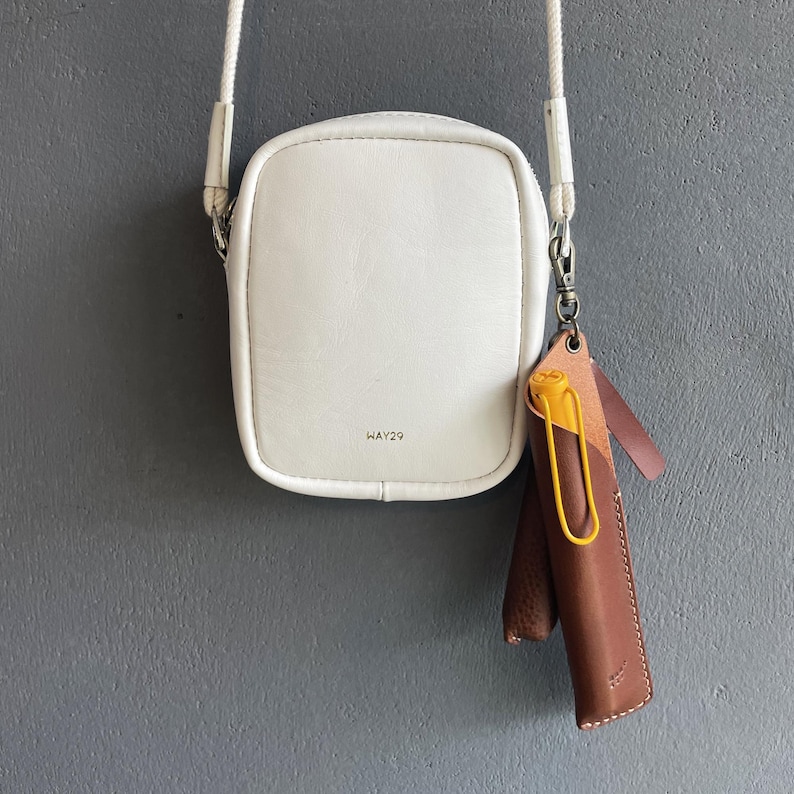 May include: A white rectangular crossbody bag with a white rope strap and the text WAY29 printed on the front. Attached to the side is a brown leather pen case containing a yellow pen, secured with a metal clip and a small leather tab.
