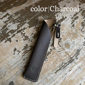 May include: A charcoal-colored leather pen sleeve featuring white stitching, a metal lobster clasp, and a small leather tag with the text JUNG. The text color Charcoal is visible at the top of the image, and the sleeve has a small embossed logo.
