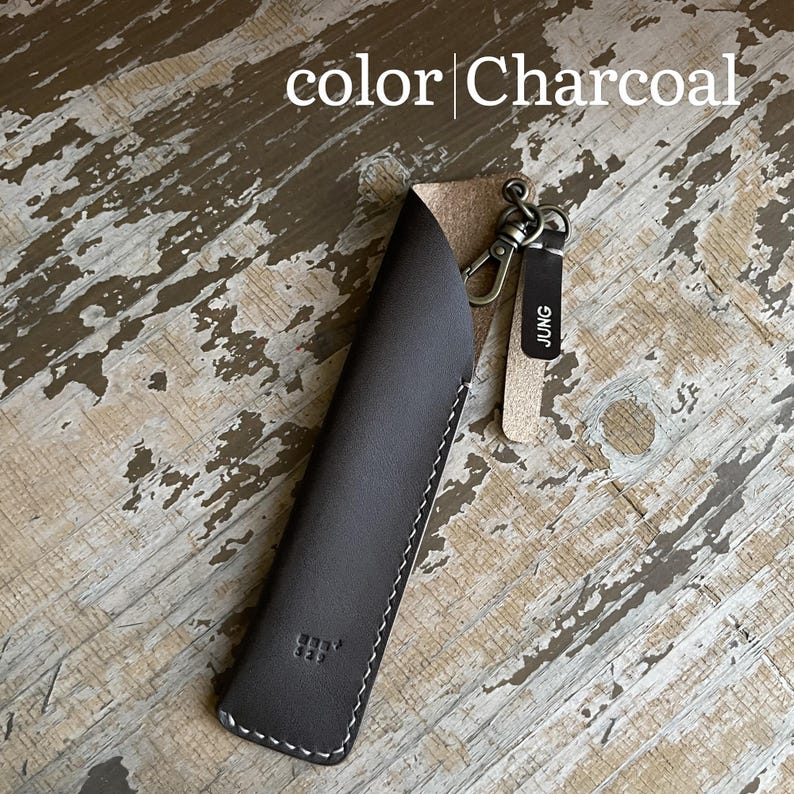 May include: A charcoal-colored leather pen sleeve featuring white stitching, a metal lobster clasp, and a small leather tag with the text JUNG. The text color Charcoal is visible at the top of the image, and the sleeve has a small embossed logo.