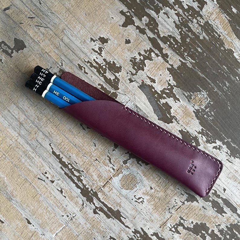 May include: A deep purple leather pencil case with white stitching along the edge and a small embossed logo. Three blue pencils with black ends are inserted into the case, showing the text 3H and 4H printed on the black ferrules.
