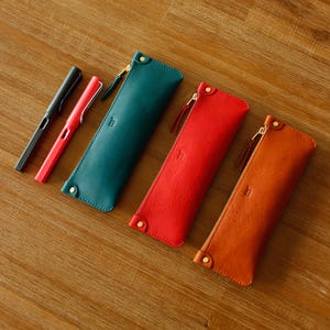 May include: Four leather pen cases in teal, red, and orange are displayed, each with a gold zip and a small stitched detail. Two pens, one black and one red, are next to the teal case. The cases are on a wooden surface.