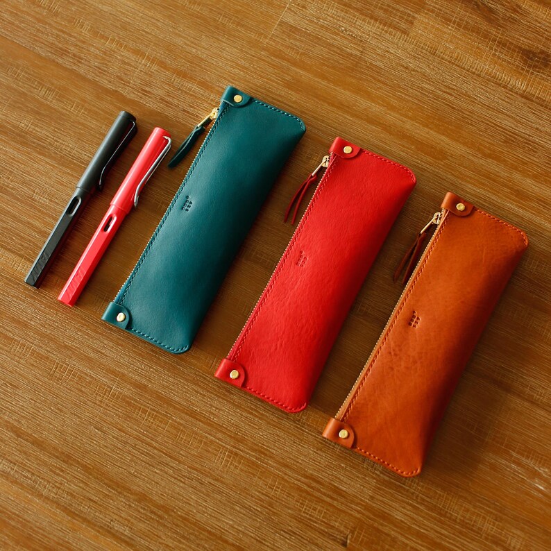 May include: Four leather pen cases in teal, red, and orange are displayed, each with a gold zip and a small stitched detail. Two pens, one black and one red, are next to the teal case. The cases are on a wooden surface.