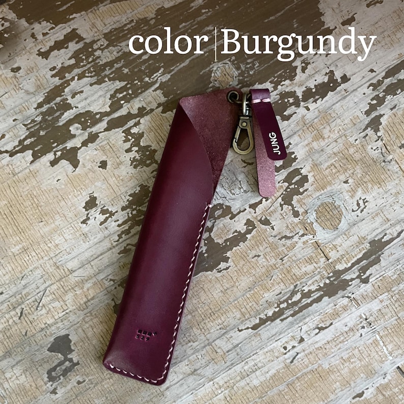 May include: A burgundy leather pen sleeve featuring white stitching, a metal lobster clasp, and a small leather tag embossed with the text JUNG. The text color | Burgundy is visible at the top of the image, and a small logo is embossed at the base.