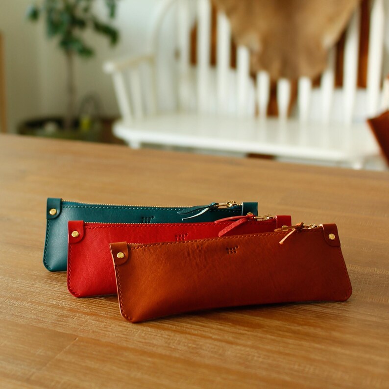May include: Three leather pencil cases in teal, red, and brown. Each case has a zip closure and a small gold-coloured rivet. The cases are rectangular and appear to be handmade, with a minimalist design.