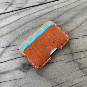 May include: A handmade, brown leather cardholder with white stitching. The cardholder has a curved cutout for easy card access and holds a blue and yellow striped card. The leather has a textured appearance.
