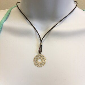 May include: A gold-toned pendant necklace with a circular, geometric design hangs from a brown leather cord.