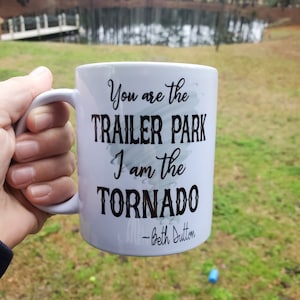 May include: White ceramic mug with the text "You are the Trailer Park, I am the Tornado - Beth Dutton" in black script. The mug is held against a backdrop of a lake and trees.
