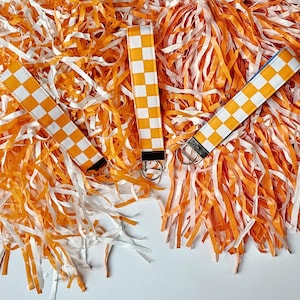 Może przedstawiać: Trzy breloczki do kluczy z pomarańczowo-białymi wzorami w szachownicę, każdy ze srebrnym metalowym kółkiem. Breloczki są umieszczone na tle pomarańczowo-białych papierowych serpentyn, tworząc żywą ekspozycję.